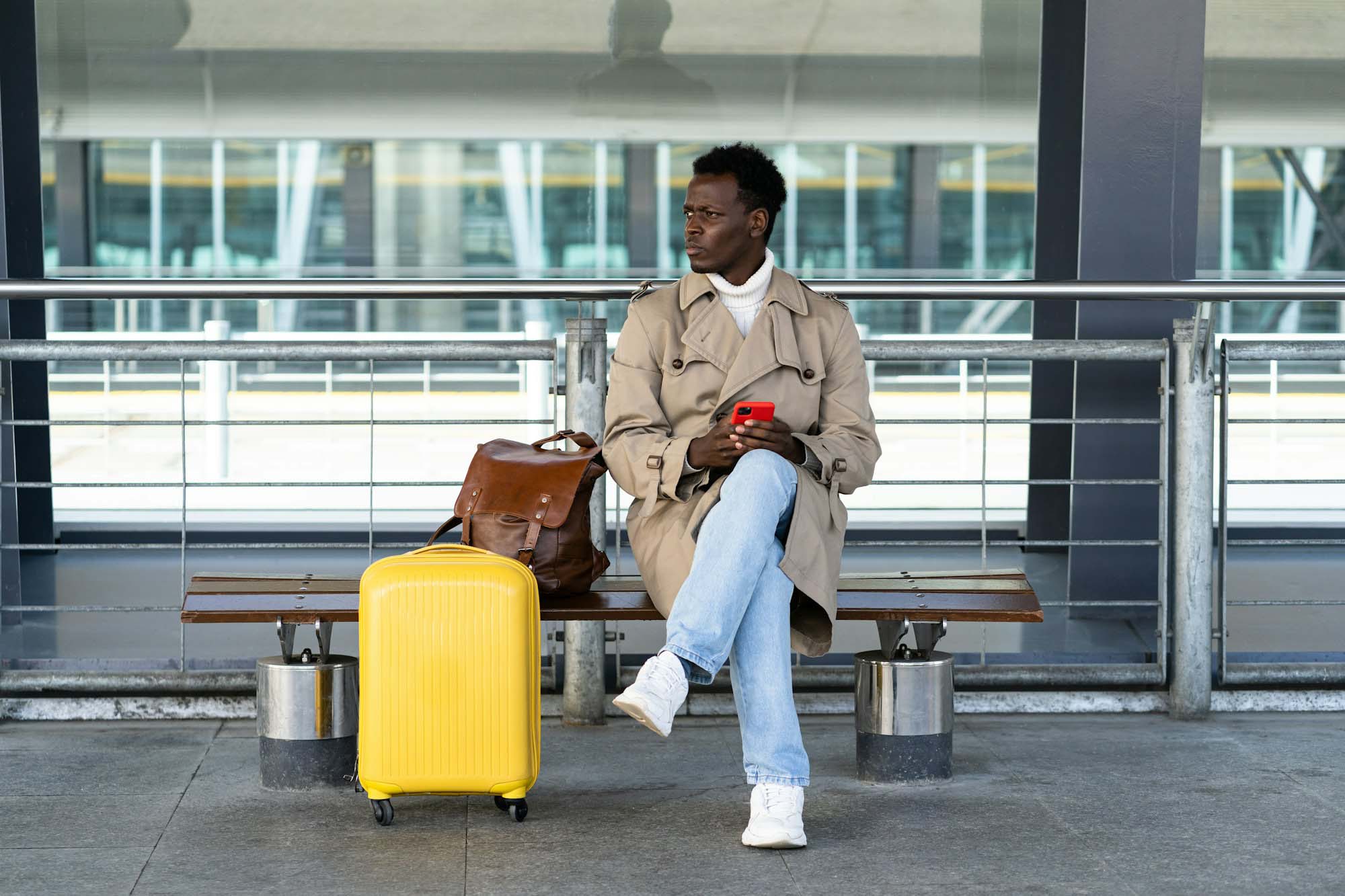 Black traveler man with suitcase sitting on bench in airport terminal, calling and looking for taxi